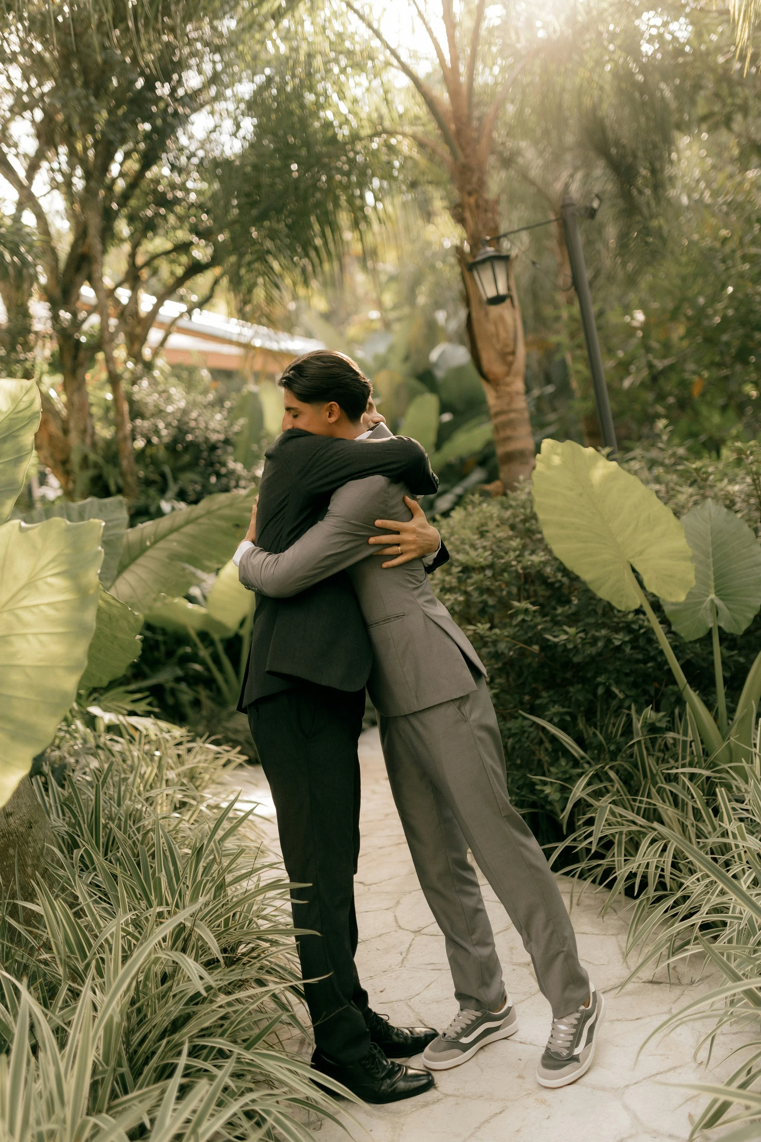 Two young men dressed in suits hugging each other in a lush, green outdoor garden area with large leaves and trees, illuminated by bright sunlight.