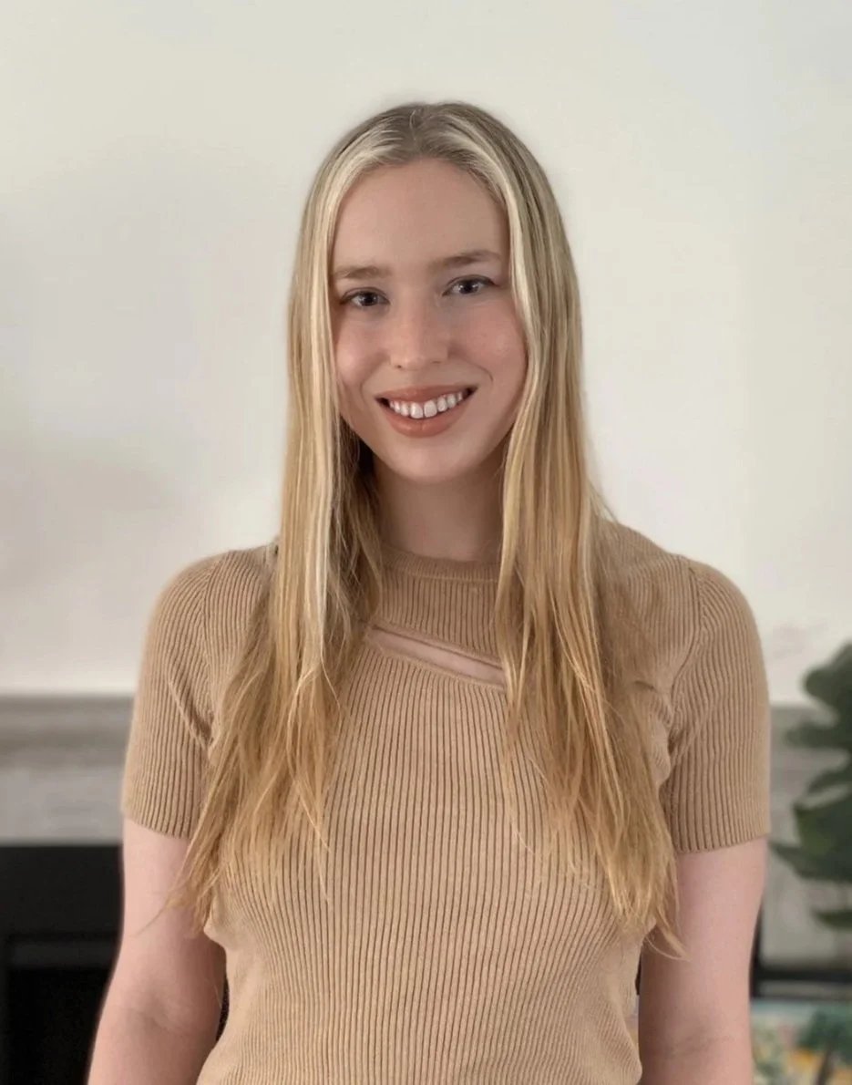 A young woman with long blonde hair wearing a tan ribbed shirt, smiling, standing in front of a white wall with a plant in the background.