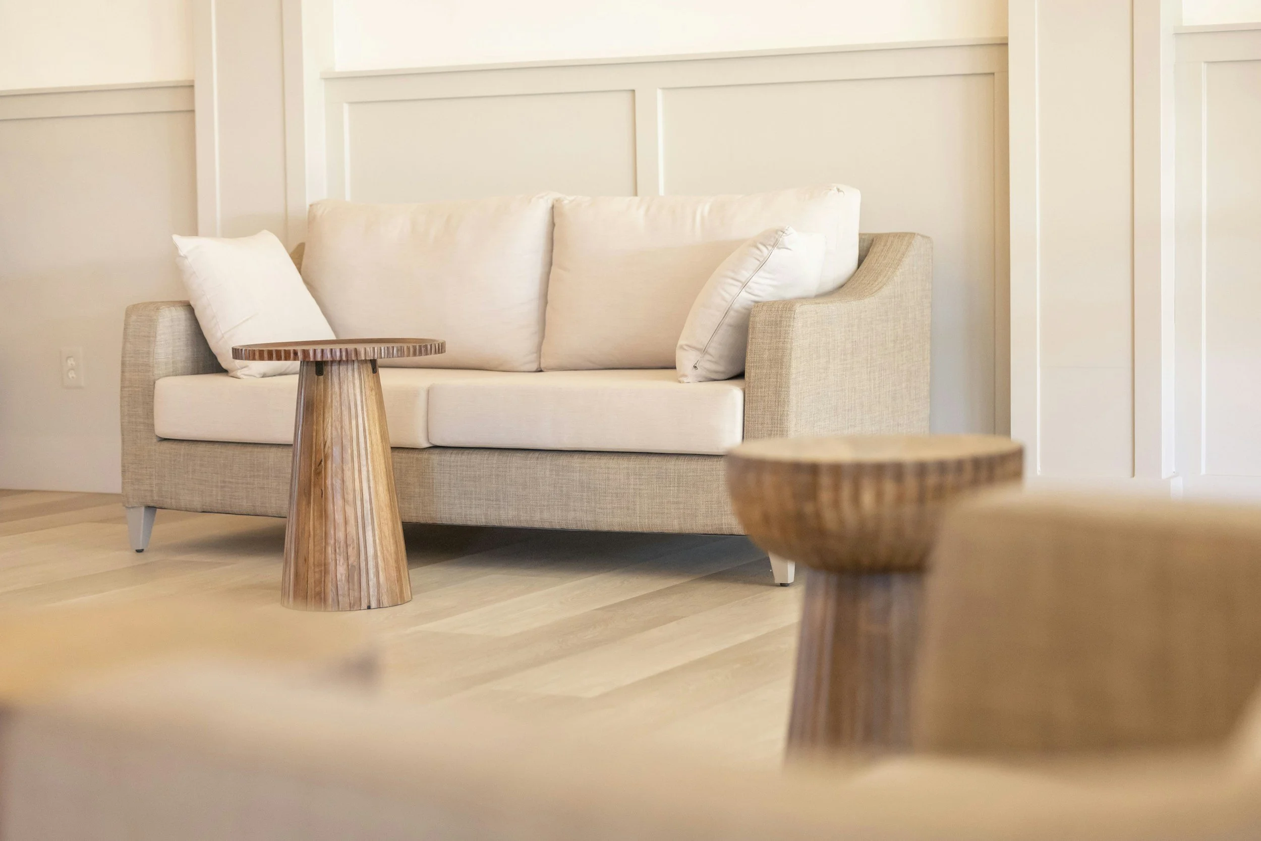 A beige sofa with white pillows in a room with light-colored wooden flooring and wall paneling. Two small round wooden tables are in front of the sofa, with one partially visible in the foreground.