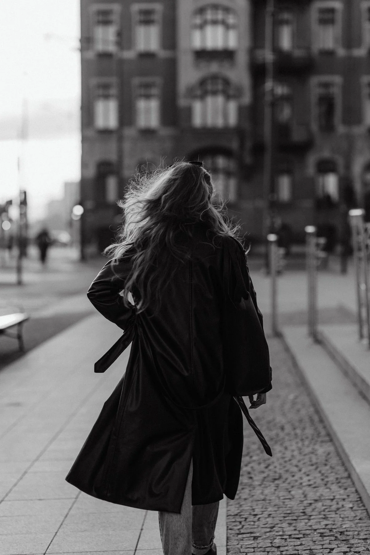 A woman with long hair wearing a black leather jacket and holding an umbrella, standing on a city street with a large building in the background. The scene is in black and white.
