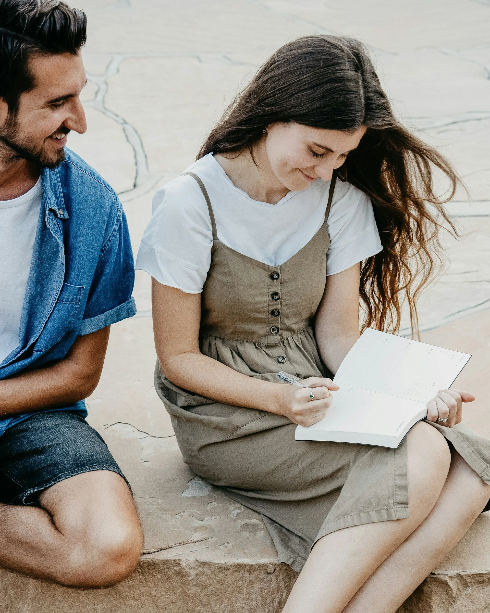 A young woman with long brown hair sitting on a rock while reading a book, with a man sitting beside her, both smiling and looking at the book.