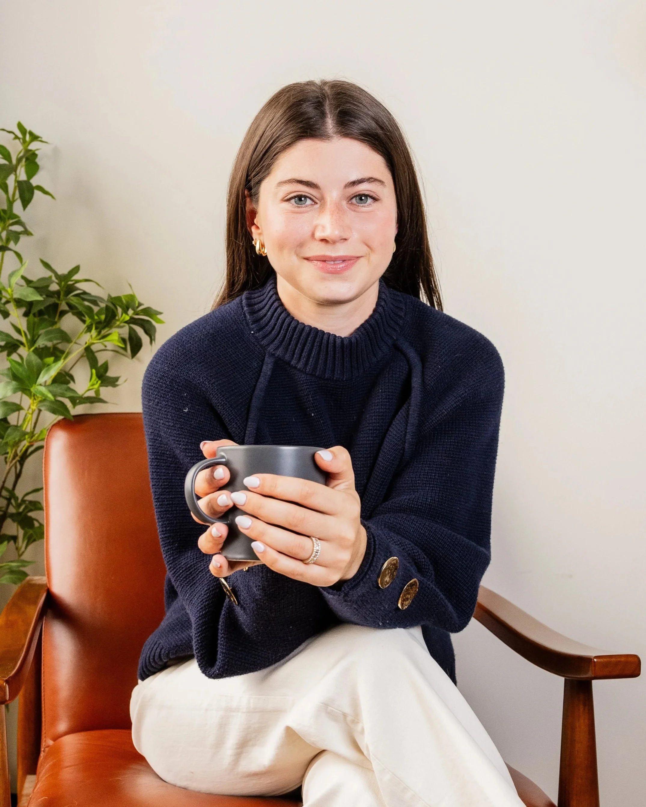 A woman with brown hair wearing a navy blue sweater and white pants sitting on a brown chair, holding a gray mug with both hands. She is smiling and looking at the camera. There is a green plant on her left and a white wall in the background.