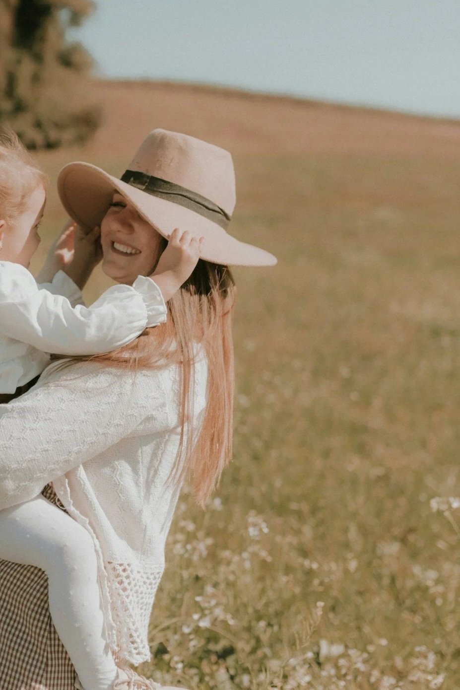 A woman with long red hair wearing a large wide-brimmed hat is playing with a young child in a field.