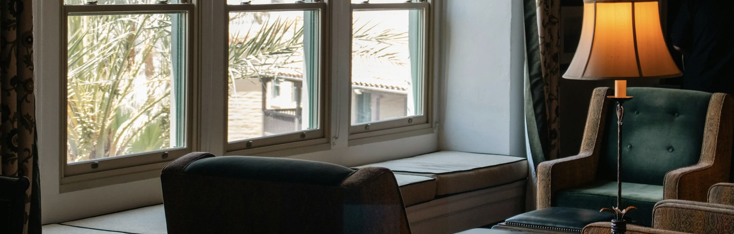 Interior of a room with large windows, a cushioned bench by the window, a green upholstered armchair, a table lamp, and a dark wall with a curtain.
