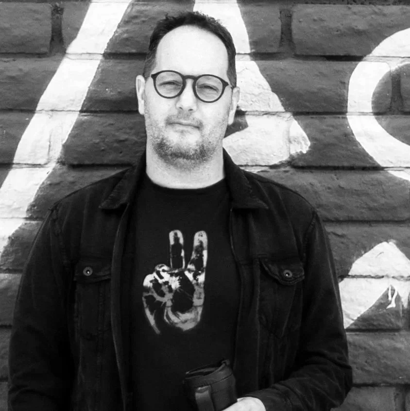 headshot picture of Larry Williams, a white man with short dark hair, wearing a black shirt with an image of a hand making a peace sign under a dark long-sleeved shirt, as he leans against a painted brick wall; photo is in black and white