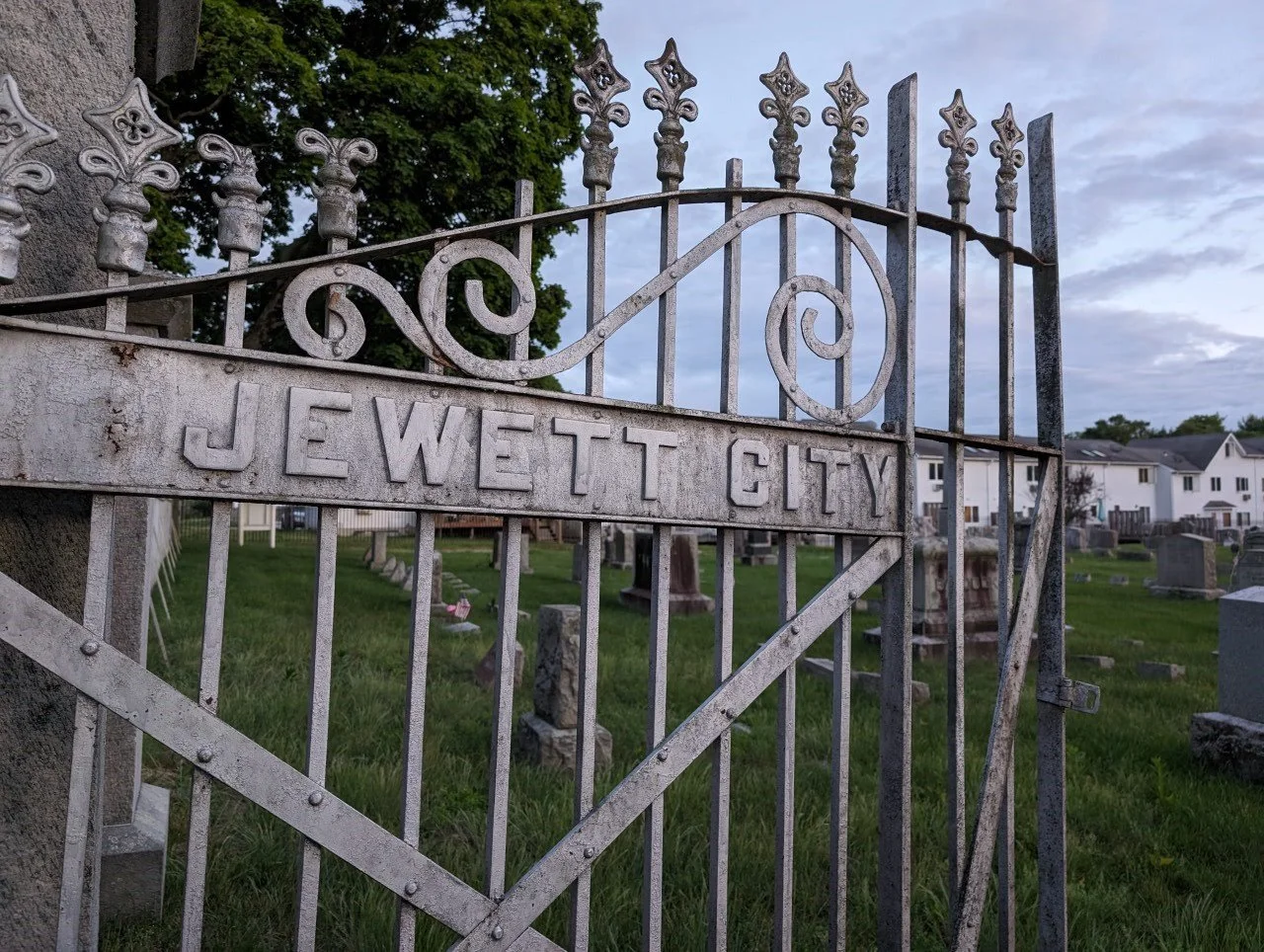 A metal gate reading "Jewett City"; behind the gate is a Victorian era cemetery.