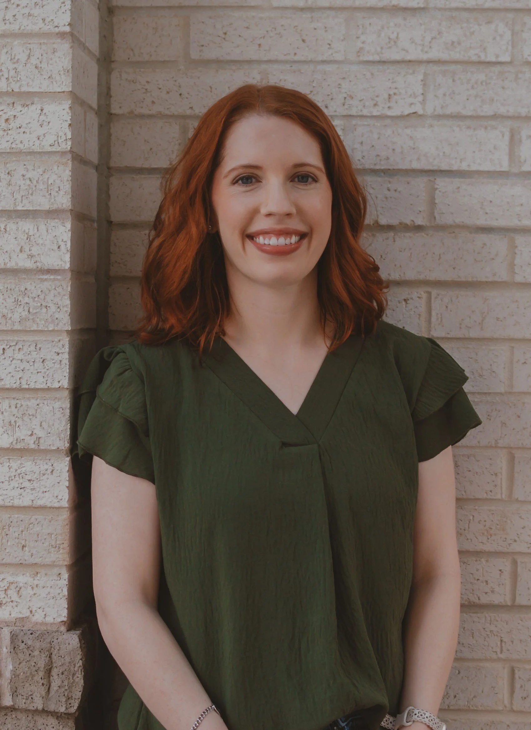 Amanda Parmley, a female therapist with red hair wearing a gree shirt & standing in front of a beige brick wall