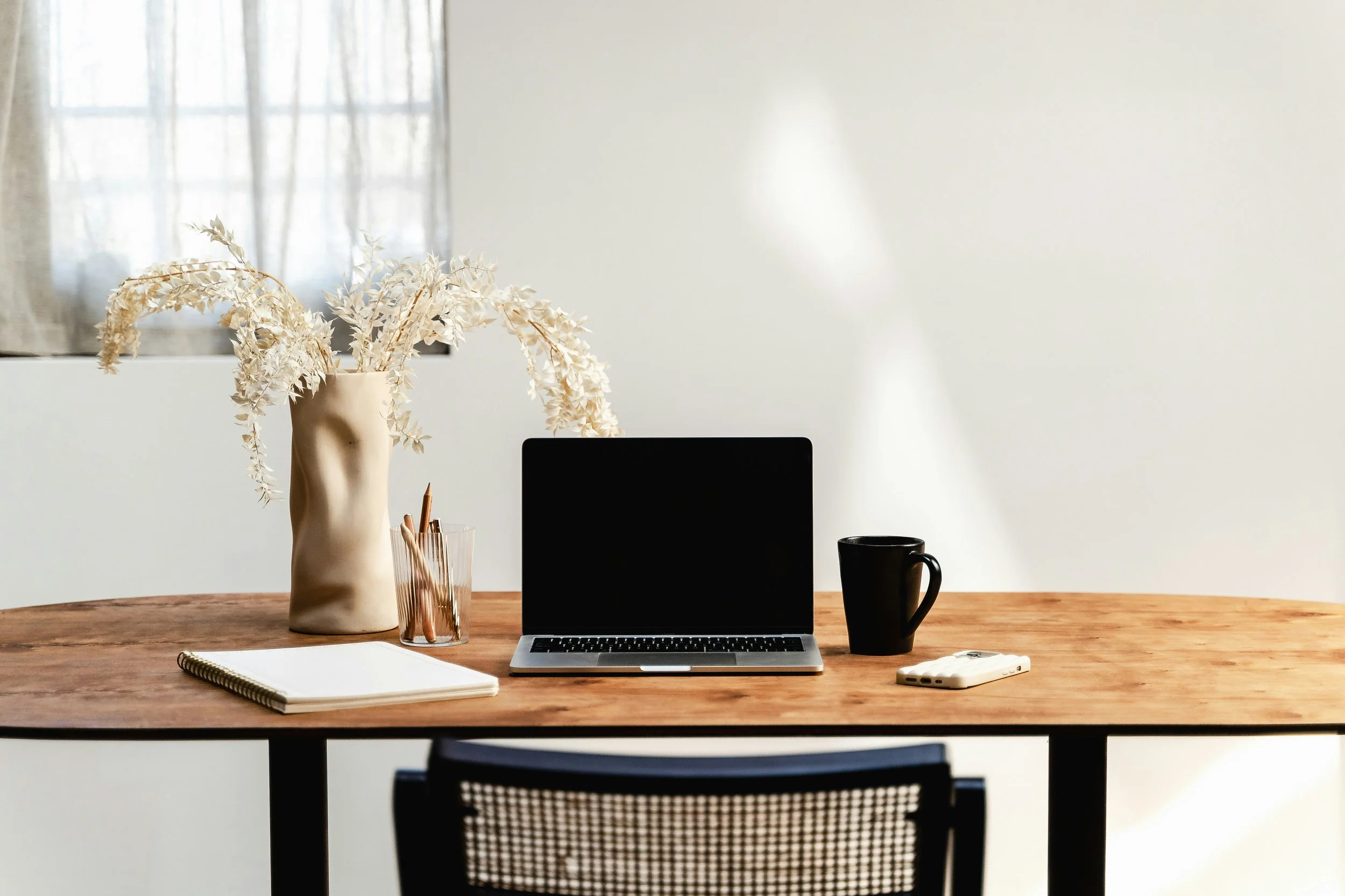 Laptop on plain wooden desk in front of grey wall with cream flowers, pencil cup, notebook, pens, black coffee mug and phone with chair in front of it.