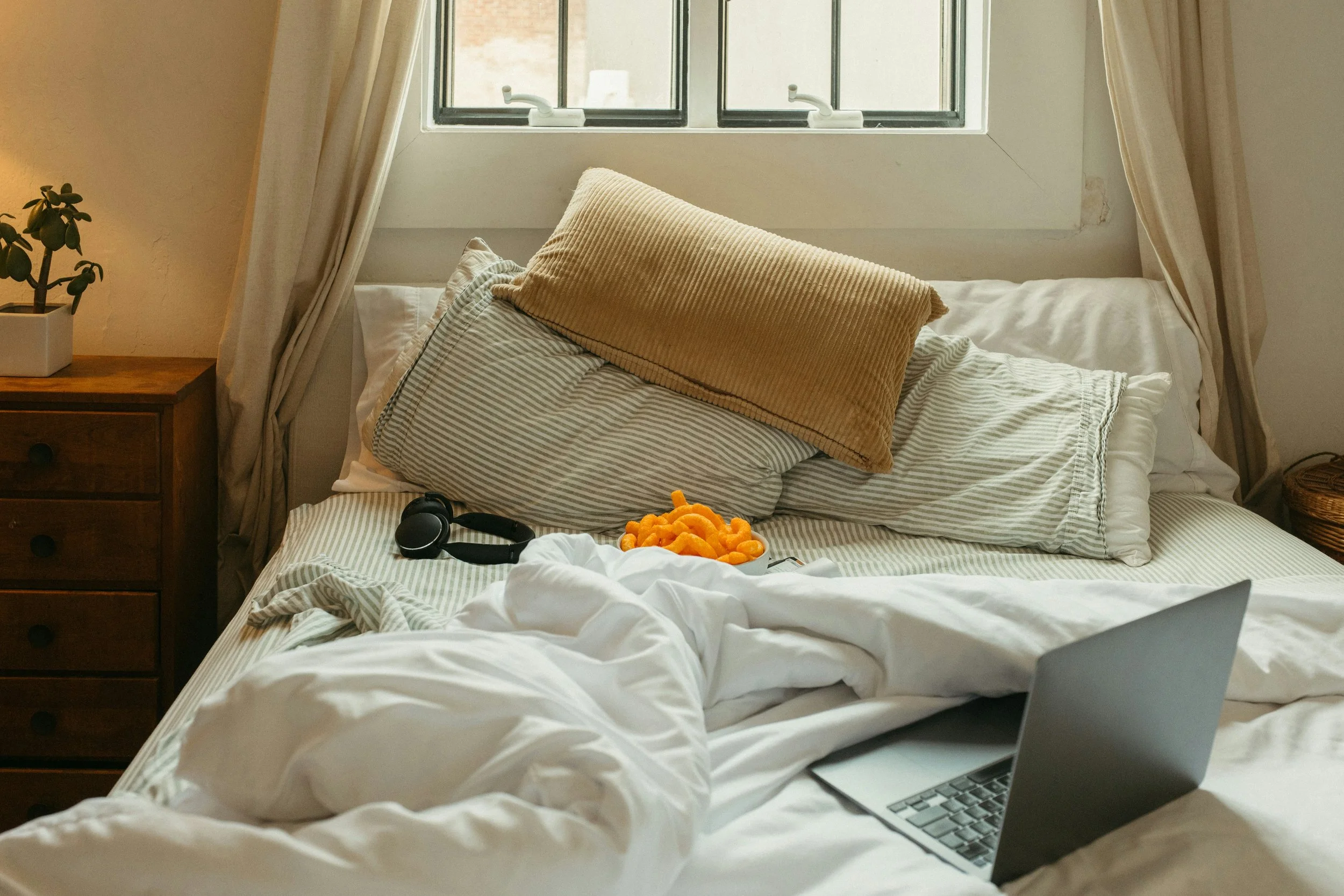 Unmade bed in front of windows with computer on the bed and headphones. Dresser besdie the bed with a small plant on it.