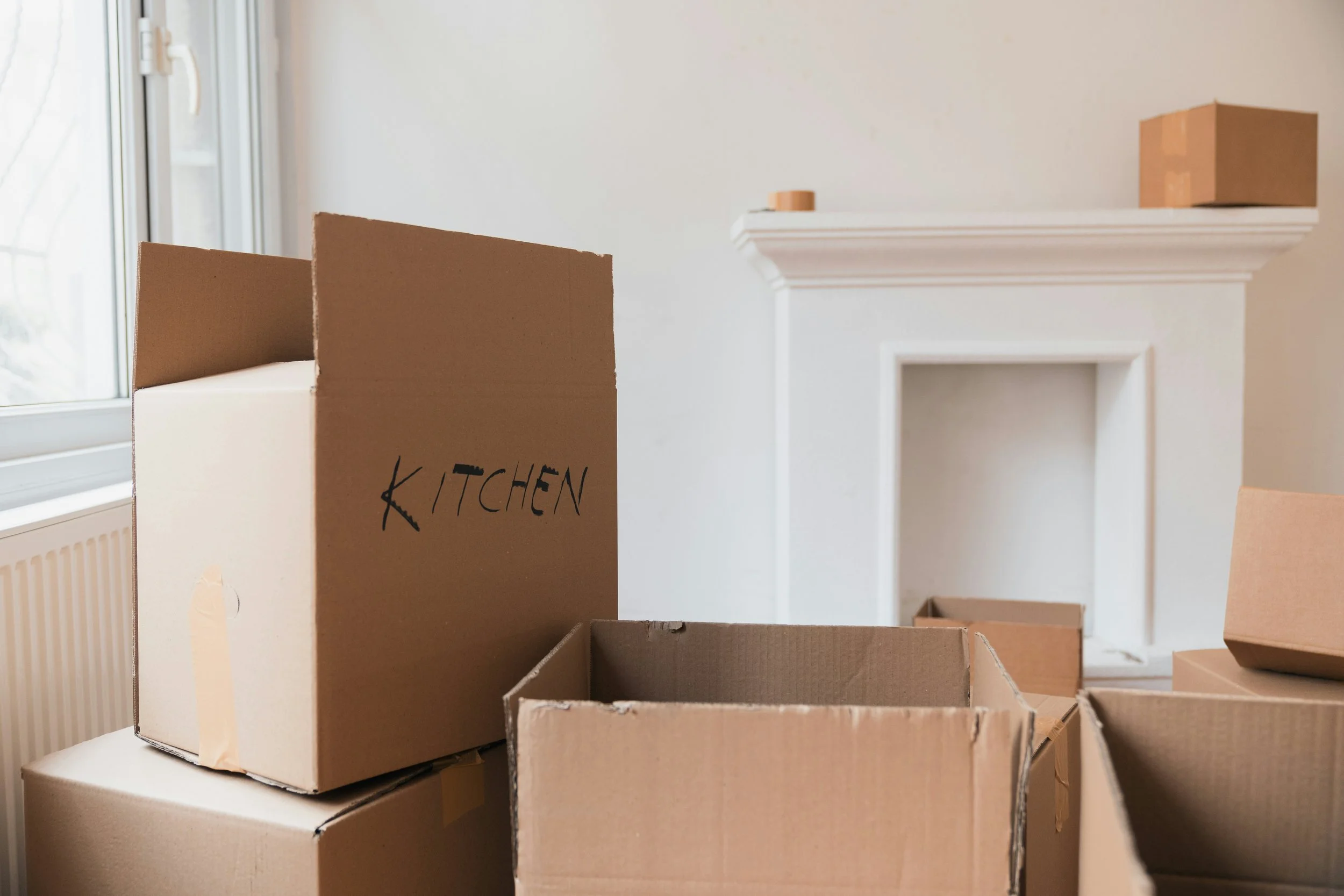 Packing boxes in front of a white wall & white fireplace. One box has "kitchen" written in black marker.