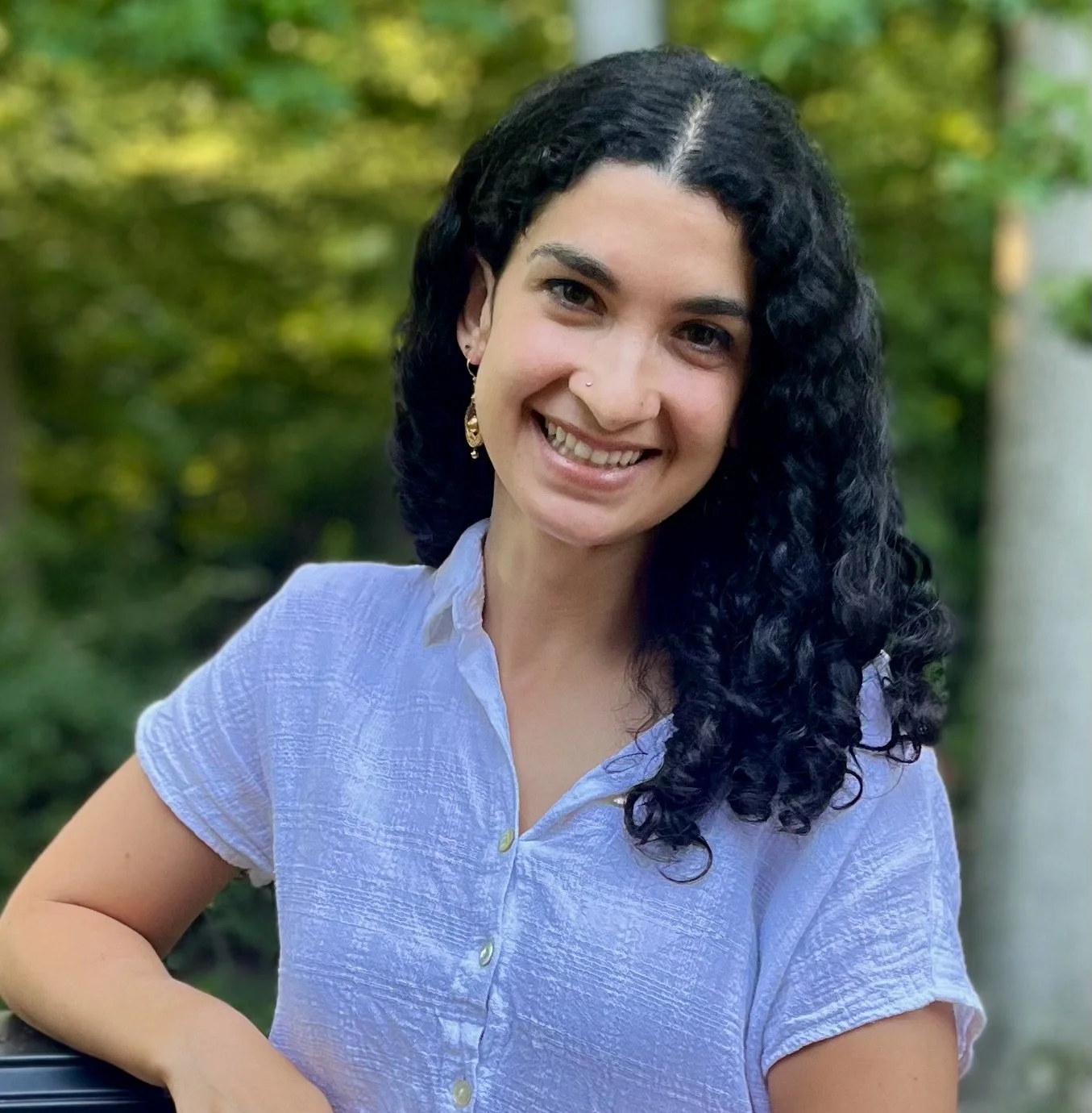 A woman with curly dark hair, wearing a light blue button-up shirt, smiling outdoors with a green, leafy background.