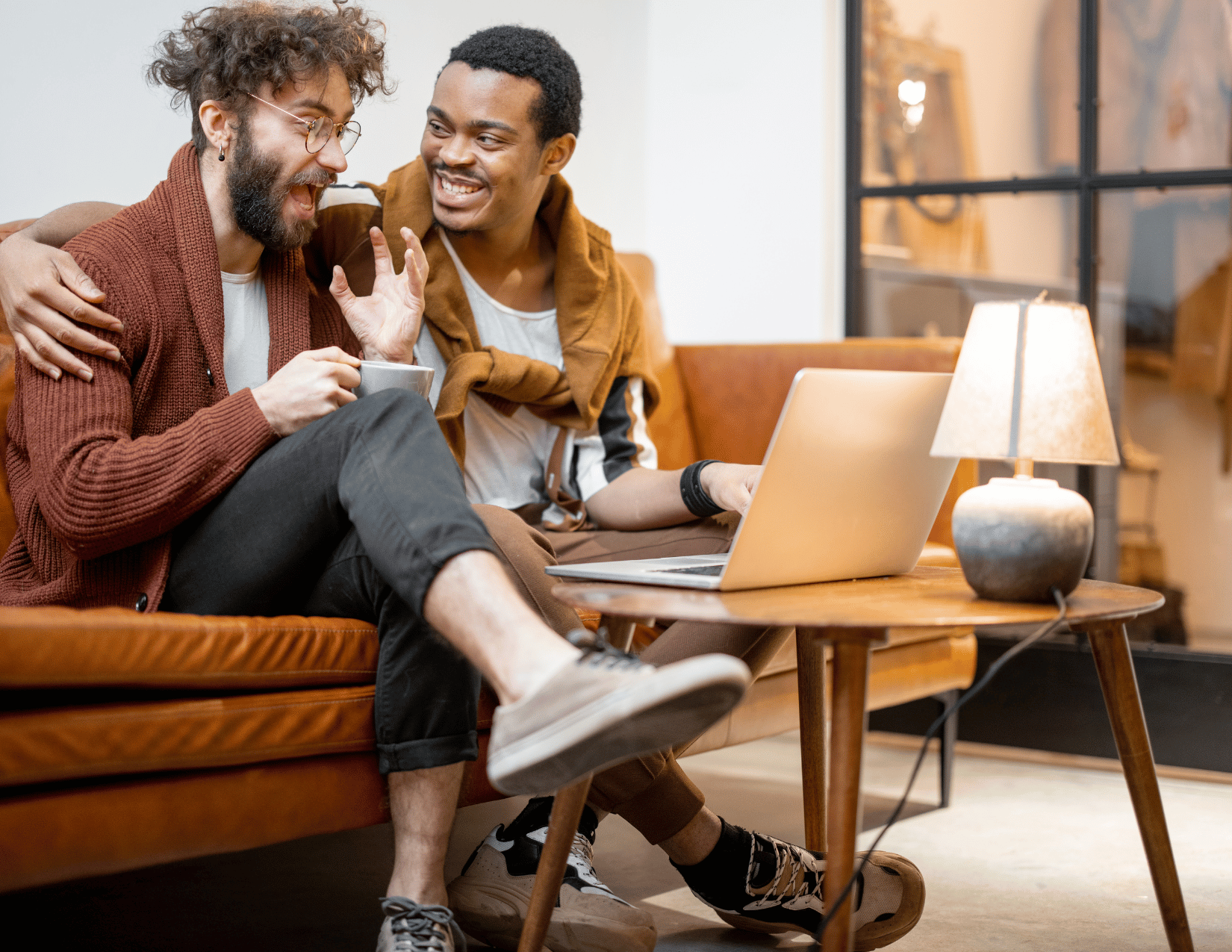 Two young men are sitting on a brown leather couch, sharing a moment of laughter and camaraderie. One man is holding a coffee mug, while the other points at a laptop in front of them. The setting appears to be a cozy, well-lit living room with a side table and a lamp.
