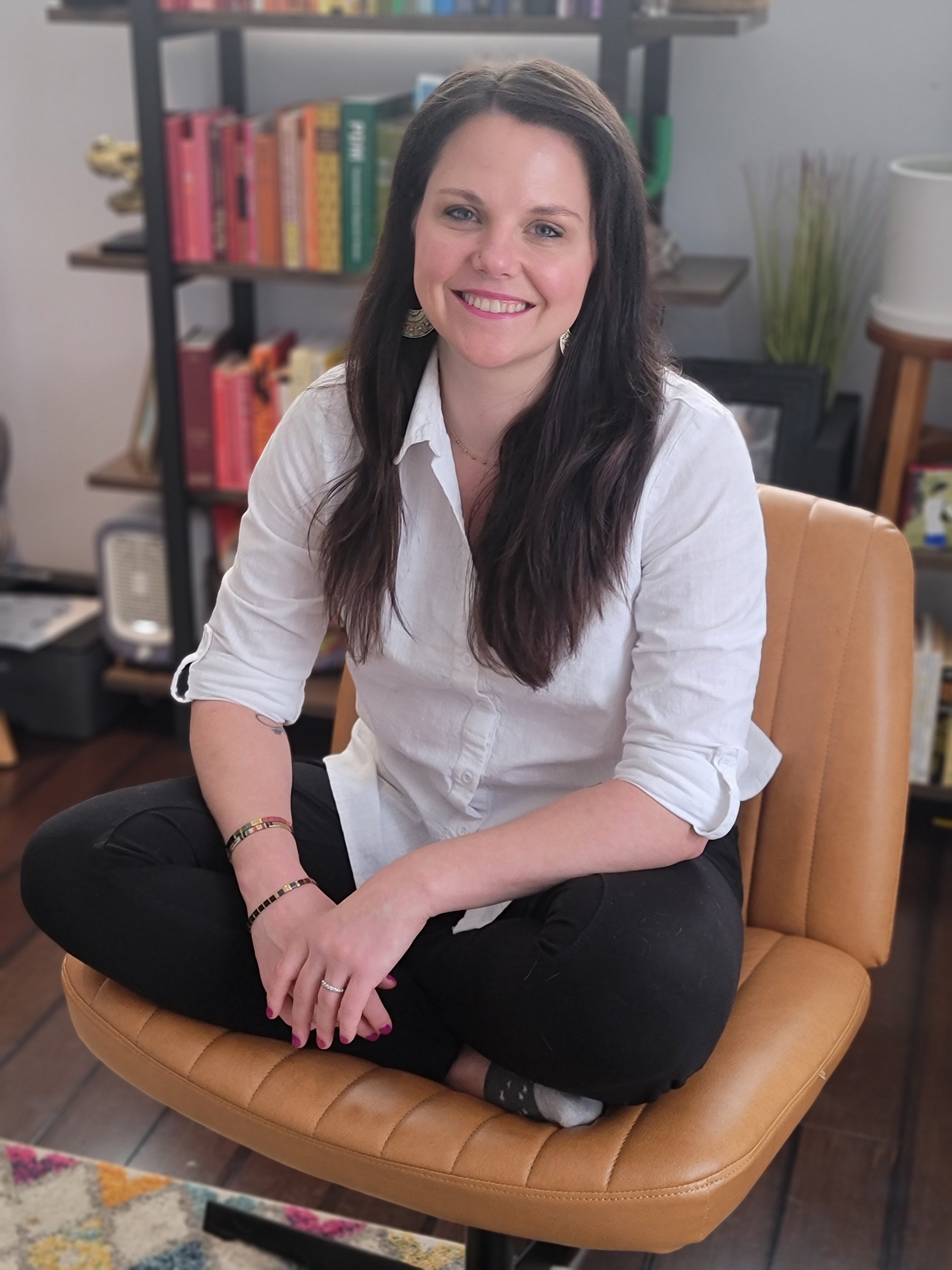 A woman with long dark hair, wearing a white button-up shirt and black pants, sitting cross-legged on a tan chair in a room with a bookshelf in the background.