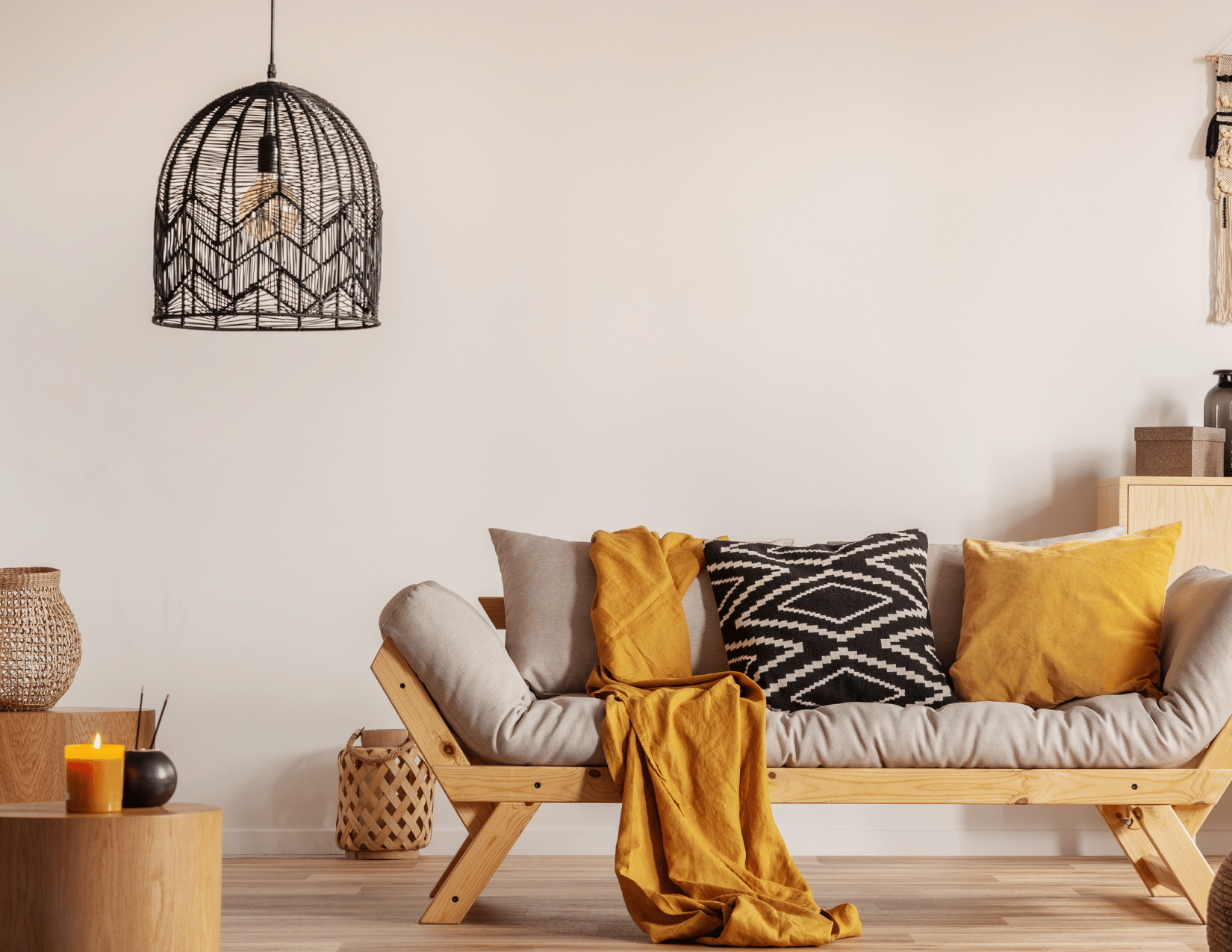A cozy living room with a modern wooden futon sofa, decorated with yellow and black patterned cushions, a yellow throw blanket, and a beige cushion. On the wooden side table, there is a lit yellow candle, a small black vase, and a larger woven basket. A black, woven pendant light hangs from the ceiling, and the room features a light-colored hardwood floor and minimal decor.
