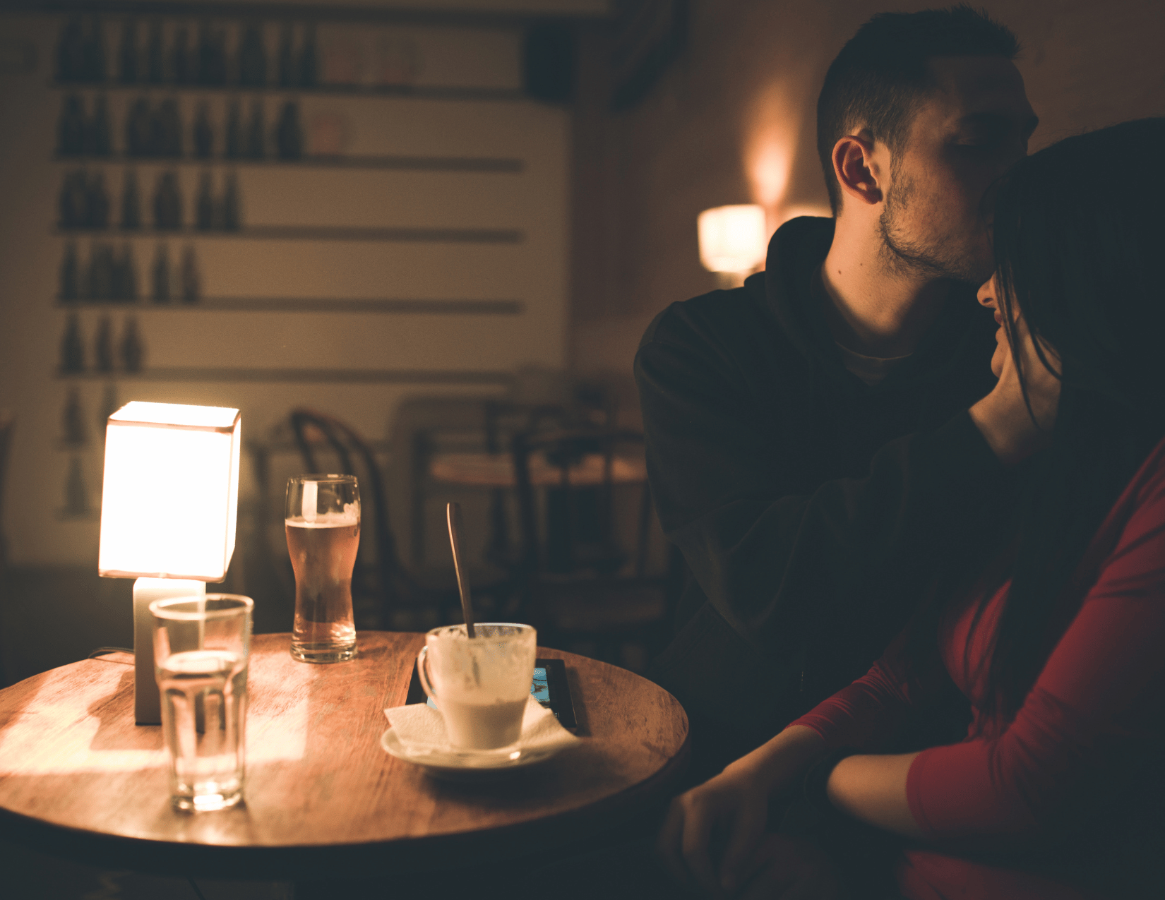 A couple sharing a kiss in a dimly lit cafe or restaurant with drinks and dessert on the table.