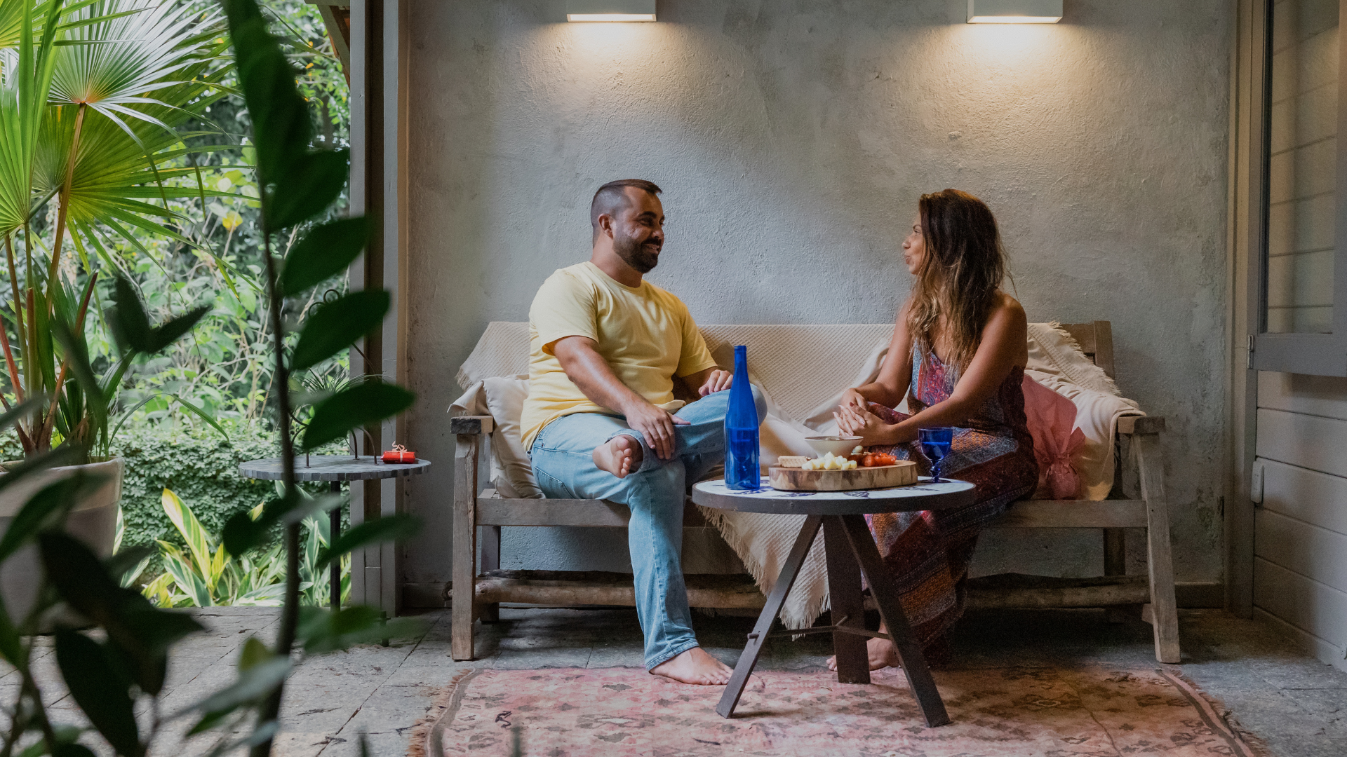 A man and woman are sitting on a wooden bench with cushions on a porch or patio, sharing a meal and talking. The man has short hair and a beard, wearing a yellow t-shirt and blue jeans, sitting cross-legged. The woman has long, wavy hair, wearing a patterned dress, with her hands resting on her lap. There are plates, a blue bottle, and wine glasses on the small round table in front of them. The setting is cozy, with greenery visible outside the open side of the porch, and warm lighting above.