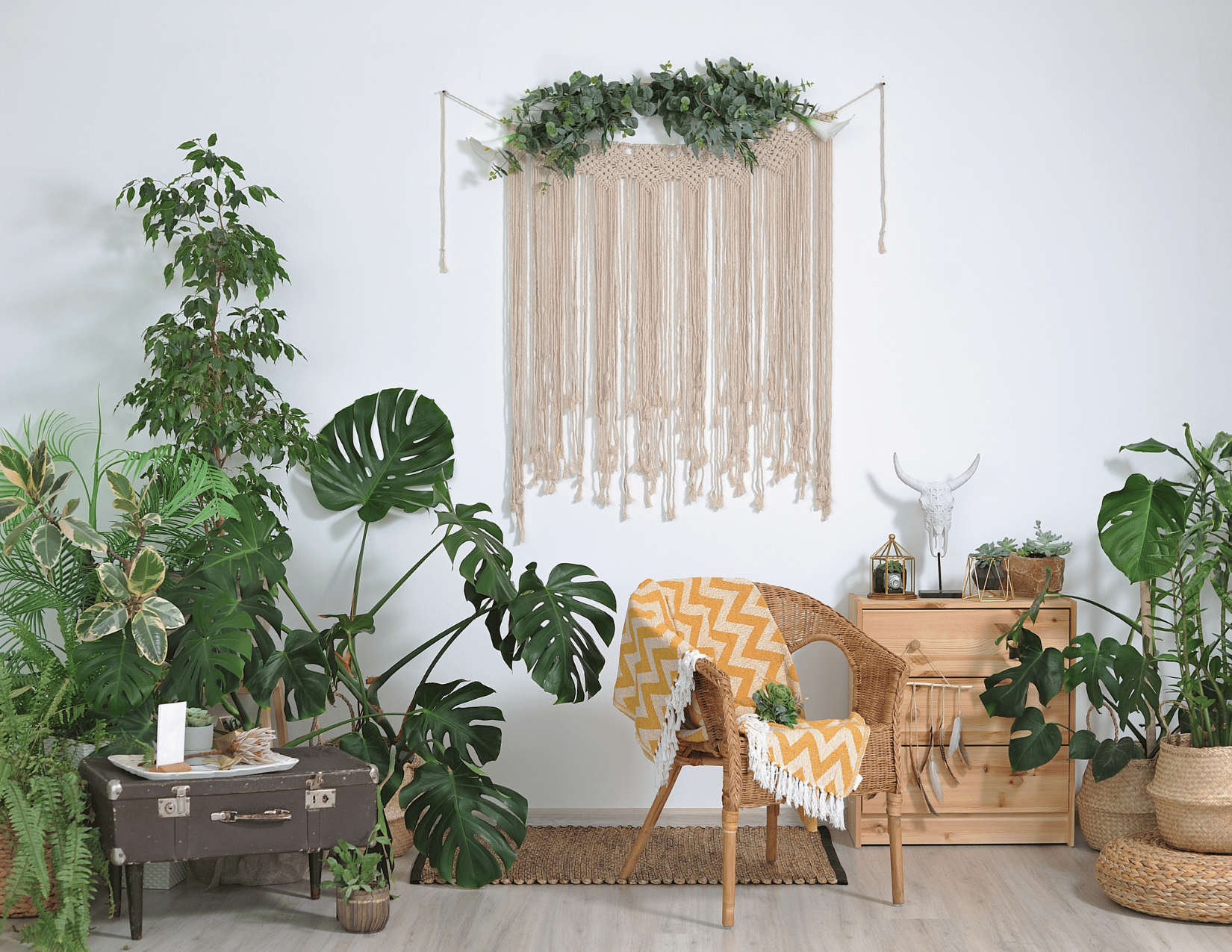 Decorative bohemian-style living room corner with wall hanging, multiple potted plants, a wicker chair with a yellow and white chevron blanket, and a wooden cabinet with decor items.