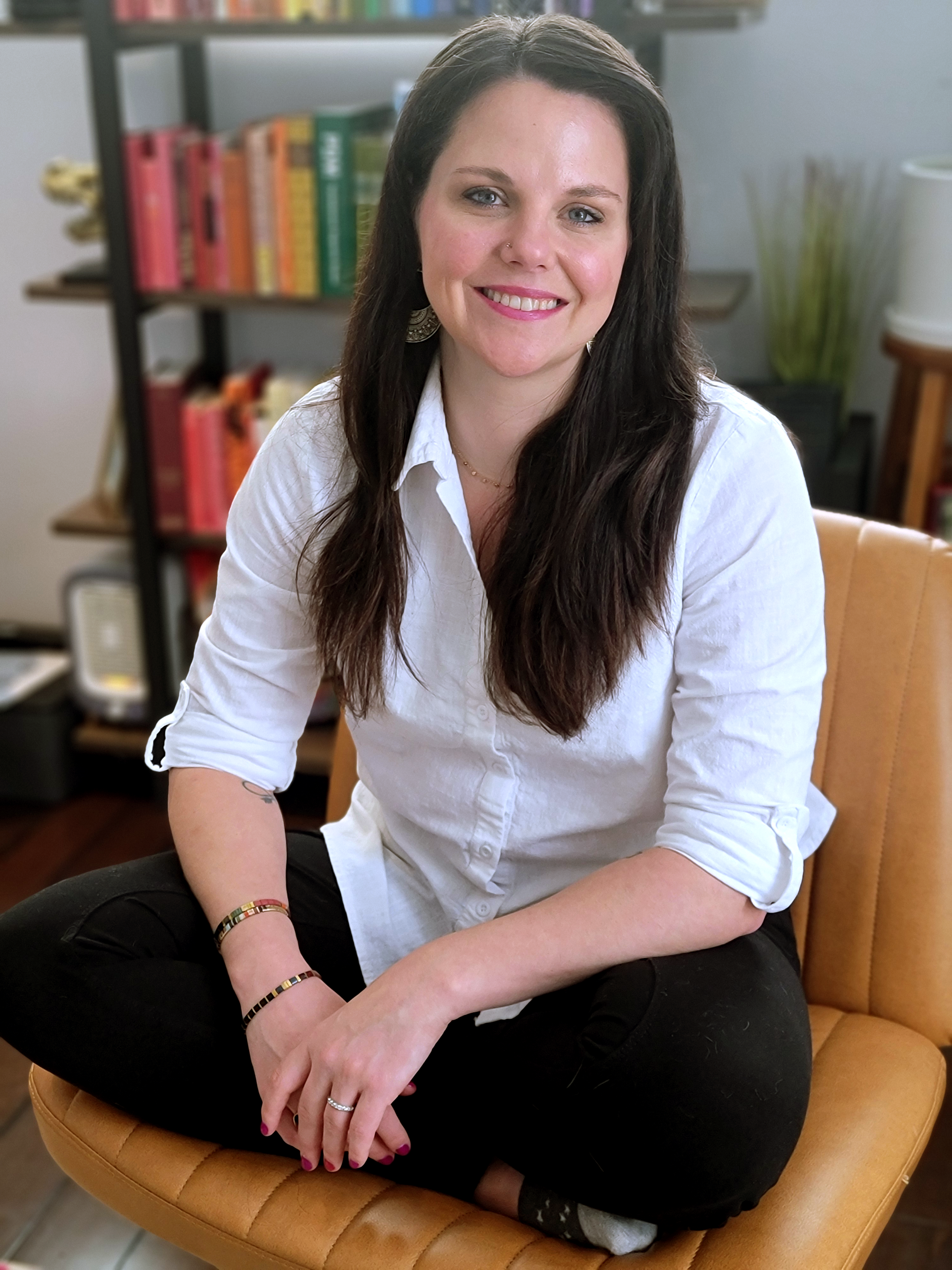 A woman with long dark hair sitting cross-legged on a tan leather chair, smiling in an office or home setting with a bookshelf and plants in the background.