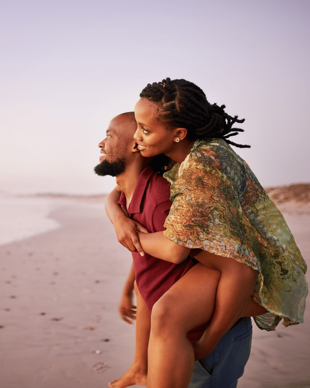 A smiling woman with dreadlocks piggyback riding on a man at the beach during sunset.
