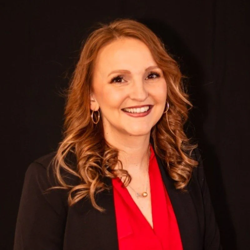A woman with long curly red hair smiling, wearing a black blazer and red top, with earrings and a necklace, standing against a black background.