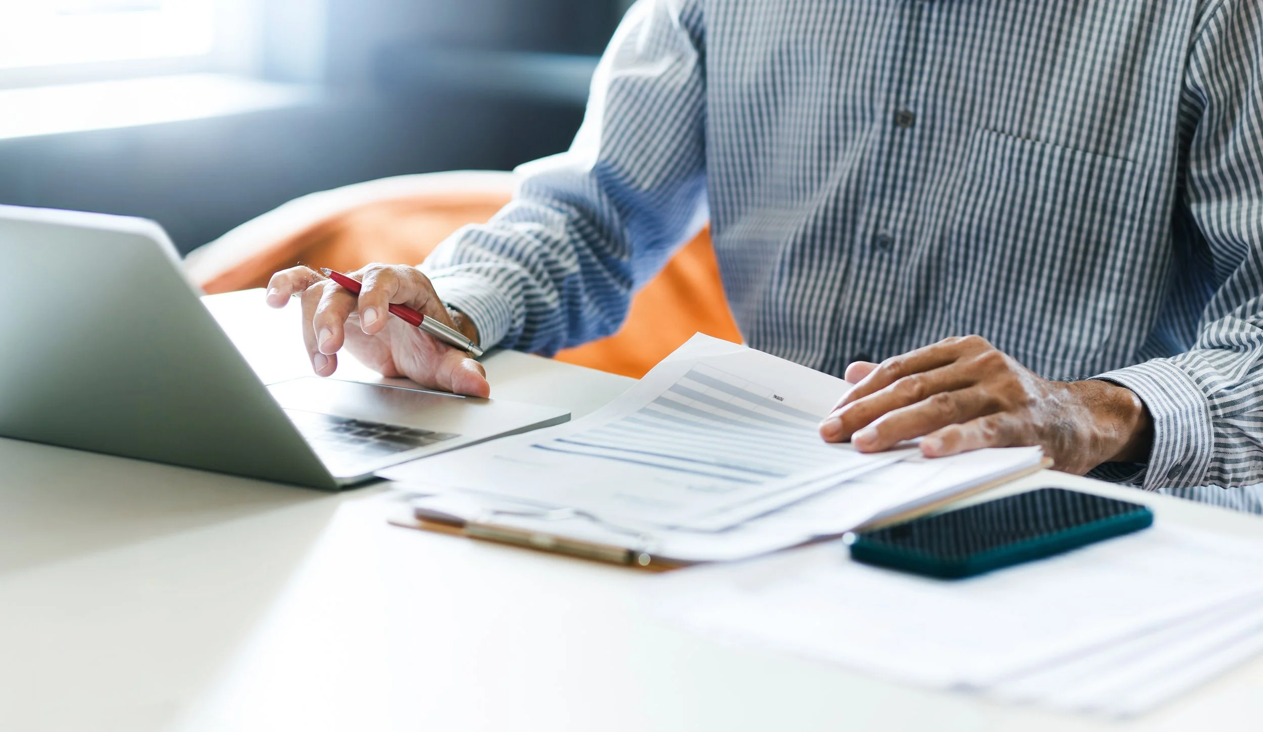 Person working at a desk with laptop, papers, and a smartphone, wearing a checkered shirt.
