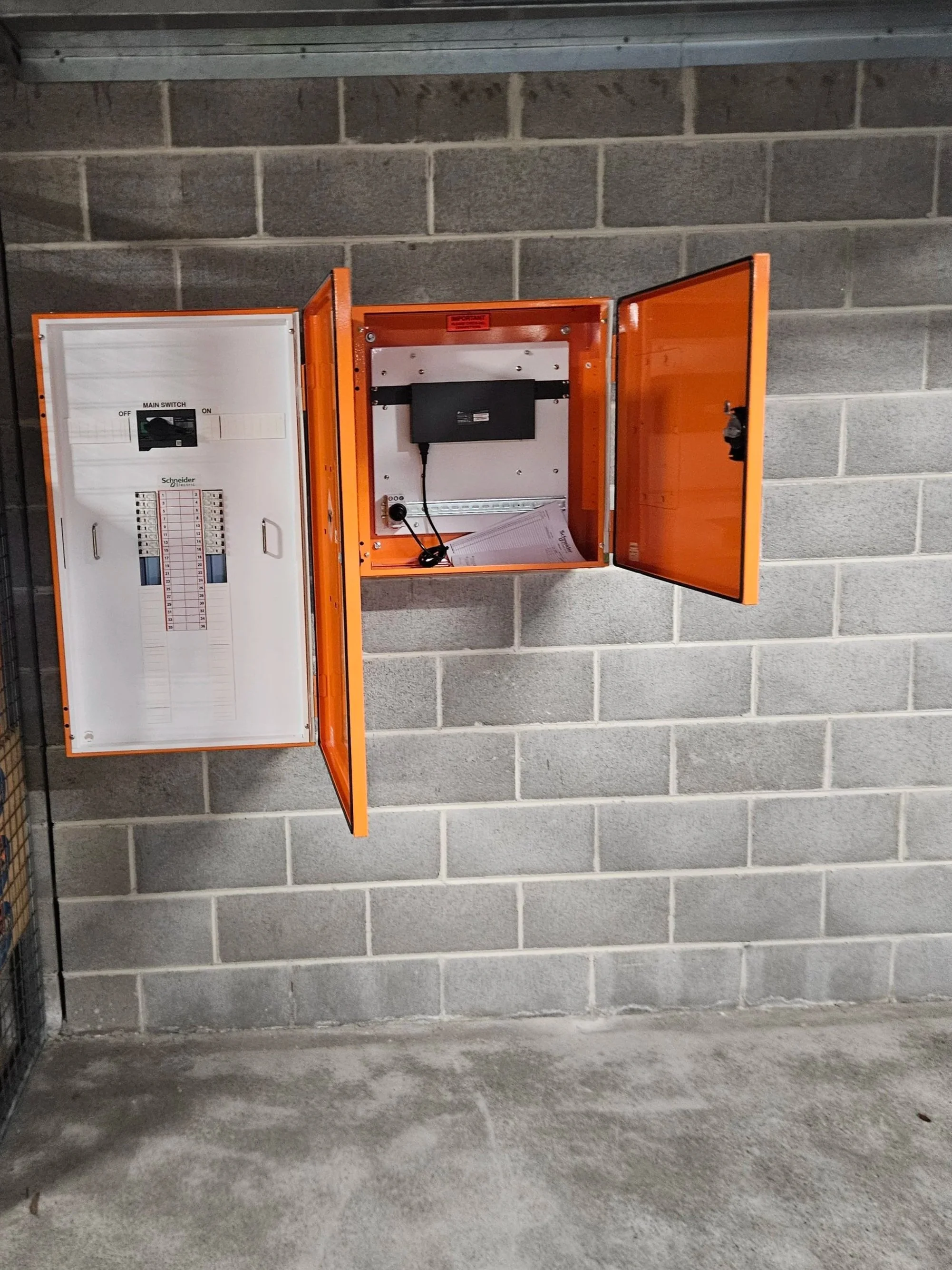 Orange electrical equipment enclosure with open door, containing a black device, some paper, and electrical components, mounted on gray cinder block wall with concrete floor.
