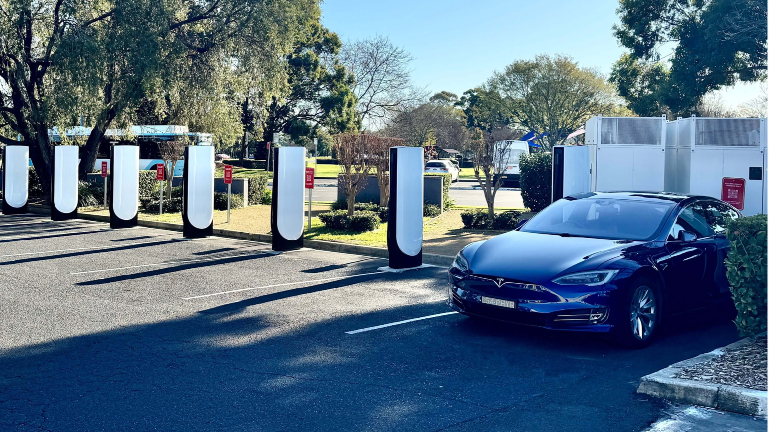 Tesla electric vehicle parked in a parking lot near charging stations, with trees and a bus in the background.