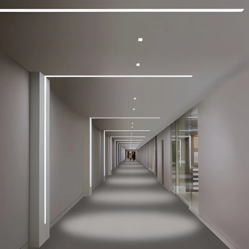 A long, modern corridor with minimalist white walls, ceiling, and floor, illuminated by linear LED lights.