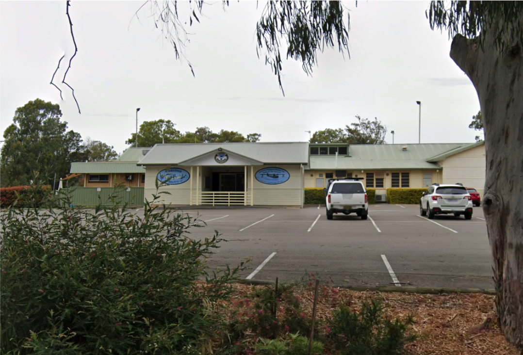 A small building with signs indicating it is an Aquatic Center, located in a parking lot with several parked cars and surrounded by trees and bushes.
