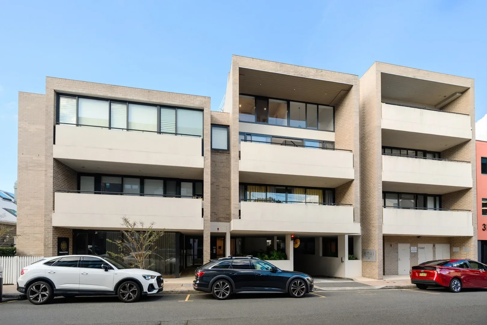 Modern multi-story apartment building with balconies and large windows, parked cars line the street in front, sky is blue with some clouds.