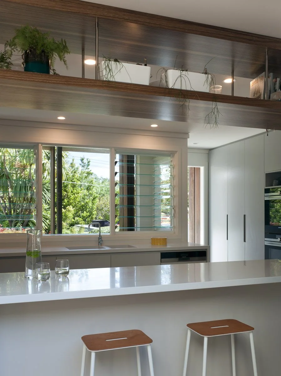 Modern kitchen interior with white countertops, bar stools, large window, and overhead shelving with plants.