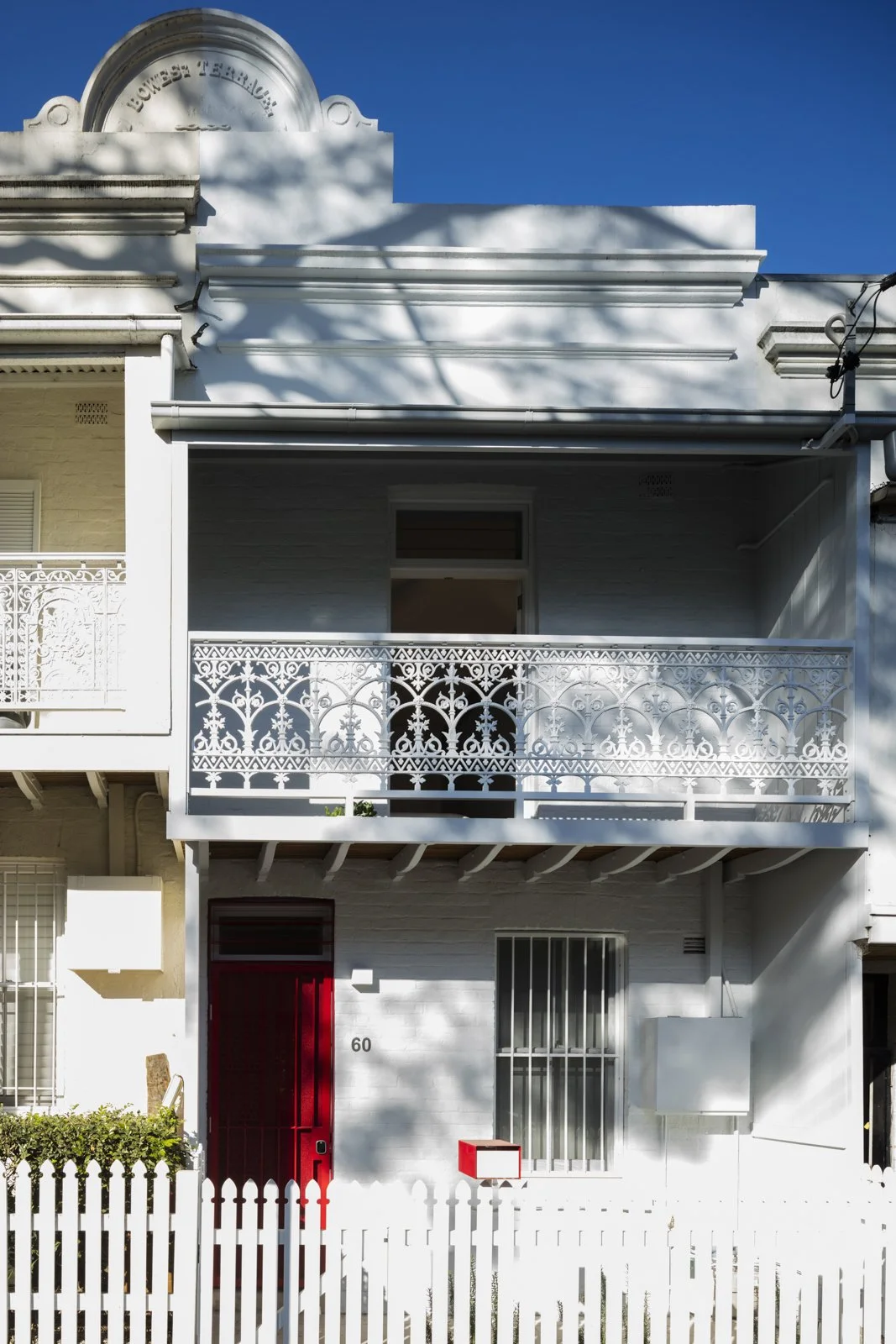 Victorian terrace house with ornate balcony, white facade, red door, red mailbox, and white picket fence.