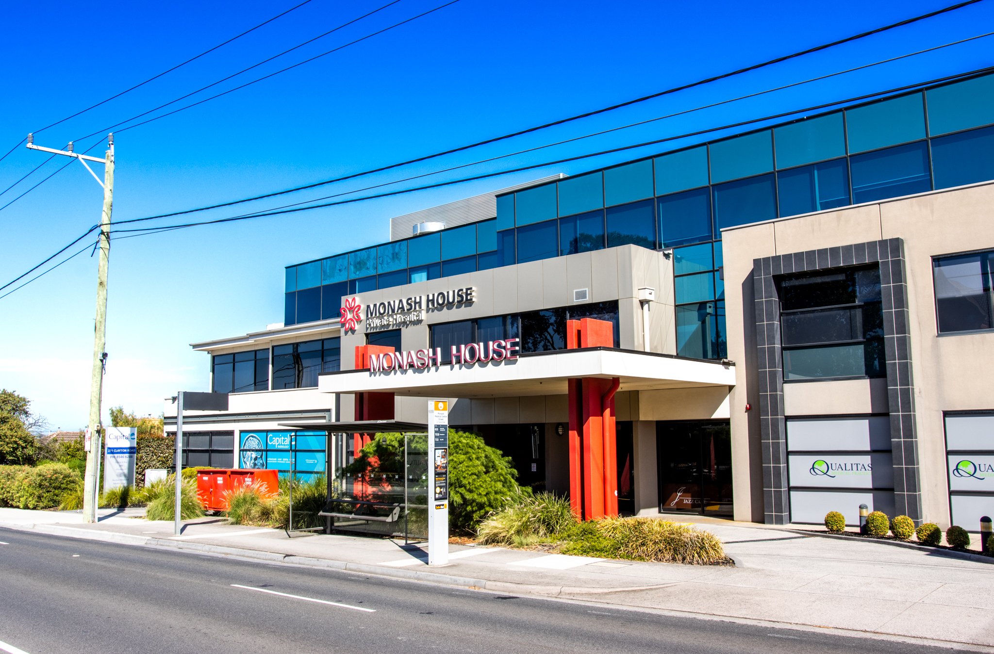 Modern commercial building with signage for Monash House and Quatlas located on a street with a bus stop, bus shelter, and bus stop sign.