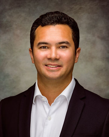 Professional portrait of a man in a white shirt and black blazer, smiling against a gray textured background.