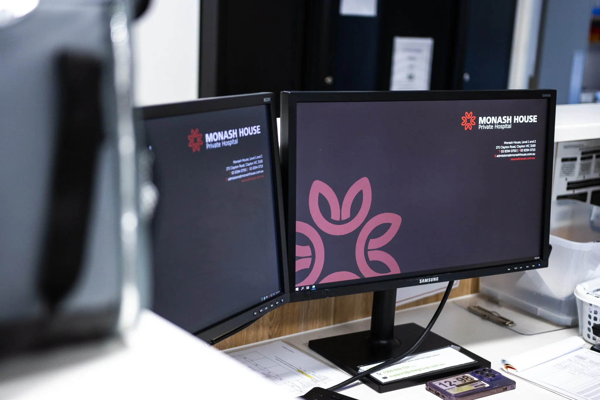 Two computer monitors displaying the Monash House Private Hospital logo and contact information on a desk in an office environment.