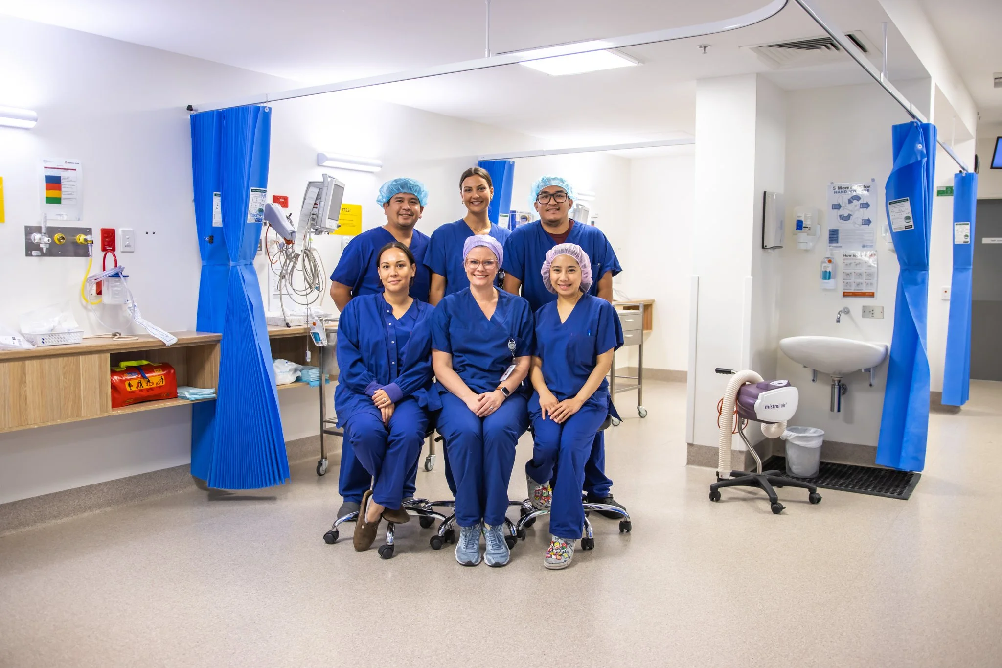 Group of six healthcare professionals in blue scrubs smiling in a hospital room with medical equipment.