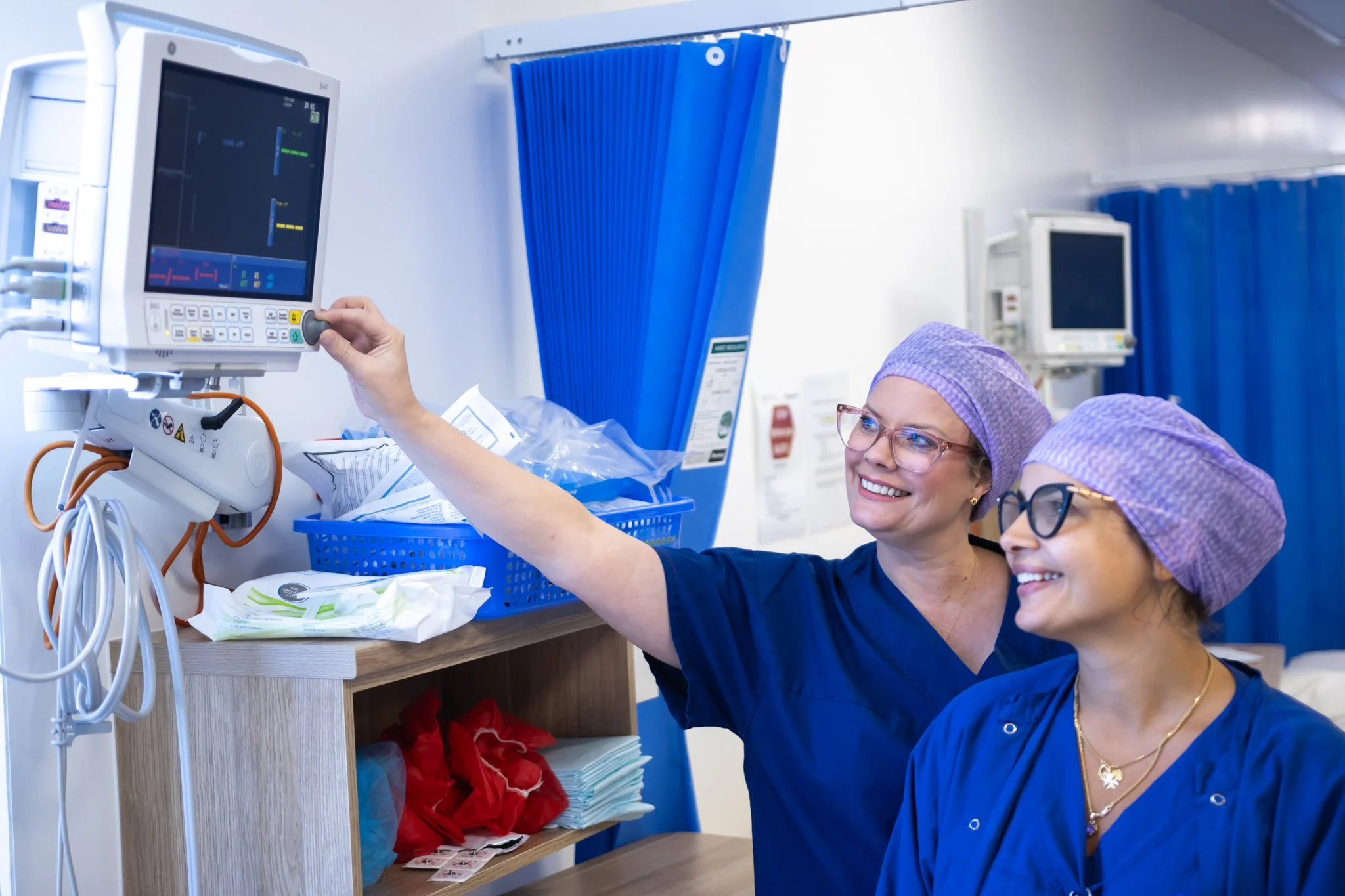 Two smiling nurses in scrubs and purple caps, one with glasses, are adjusting a monitor on a medical ventilator in a hospital room.