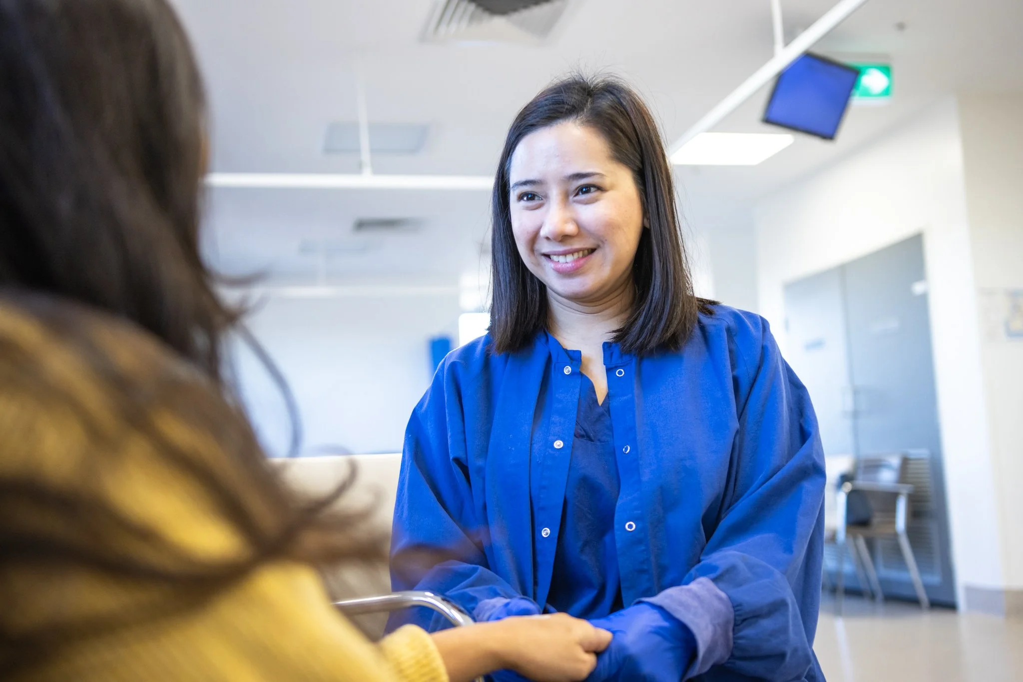 A woman in a blue uniform smiling and holding hands with another person in a waiting room or hospital setting.