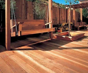 Wooden outdoor swing and platform on a wooden deck, surrounded by a wooden fence and greenery.