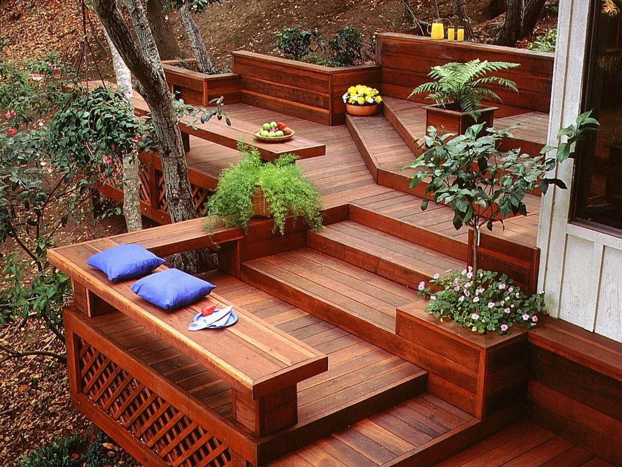 Wooden outdoor deck with built-in benches, potted plants, and yellow candles, surrounded by trees and bushes.