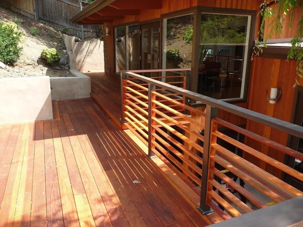 Wooden deck with railing attached to a house with large glass windows and a sliding door, surrounded by landscaping with rocks and plants.