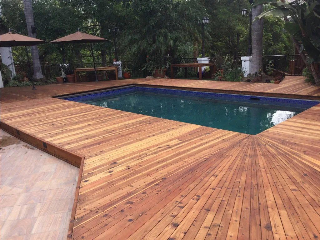 Backyard pool surrounded by a wooden deck with outdoor seating, umbrellas, and lush greenery.