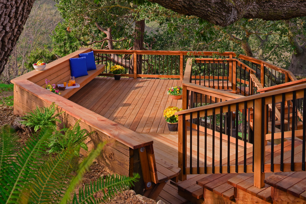 Wooden outdoor deck with stairs and railing, decorated with colorful flowers and cushions, surrounded by leafy trees.