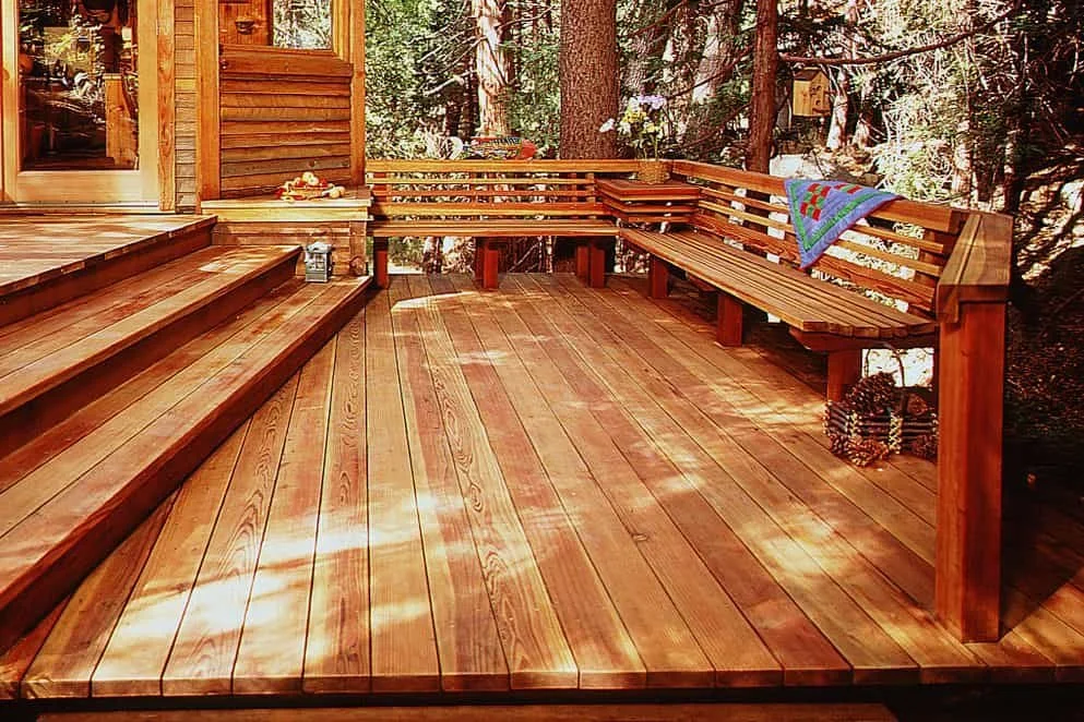 Wooden deck with built-in bench seating, surrounded by trees and a cabin, featuring a colorful throw blanket on the bench and some pinecones on the floor.