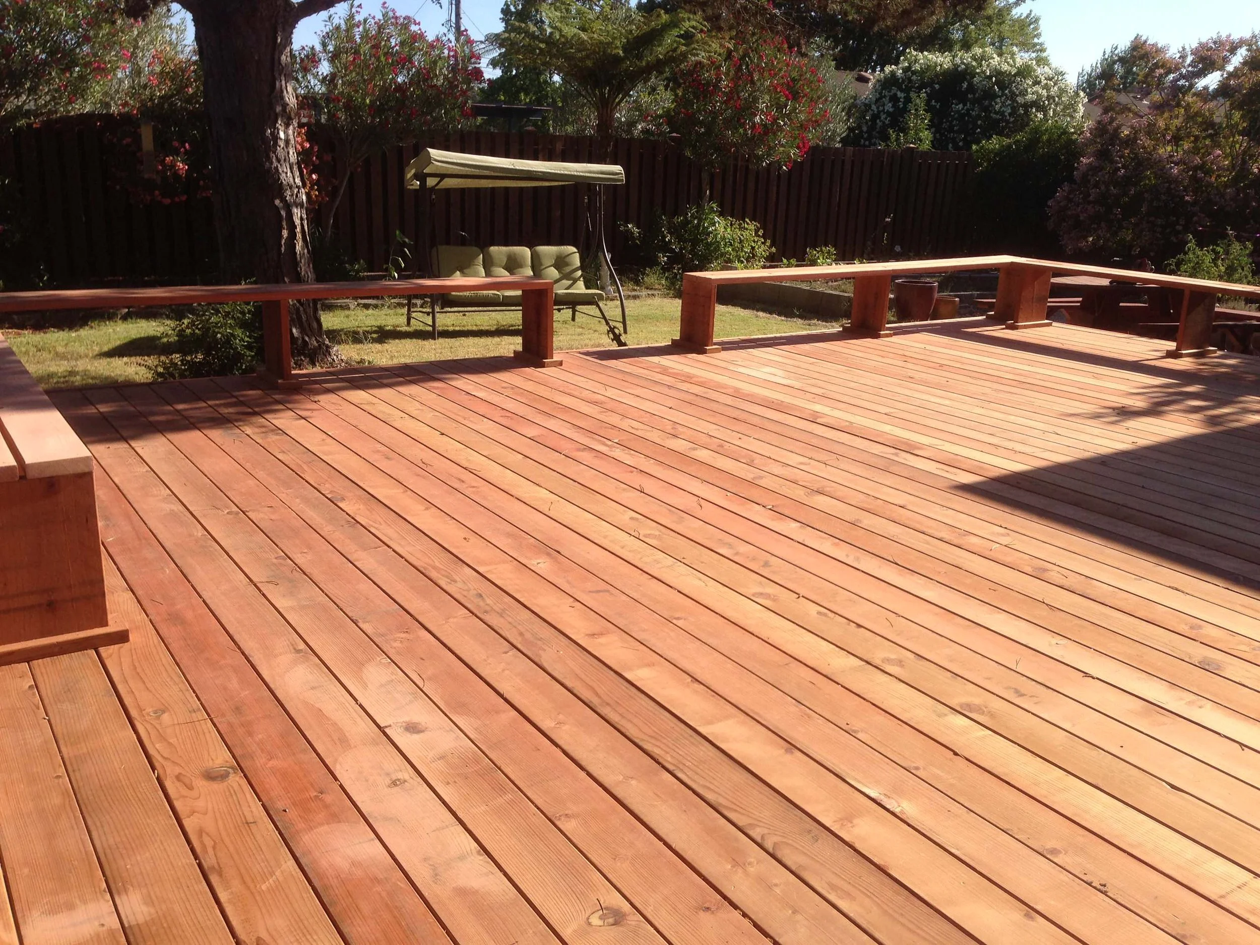Wooden deck with a garden and flowering bushes in background, a swing bench under a canopy, and a shaded area on the right.