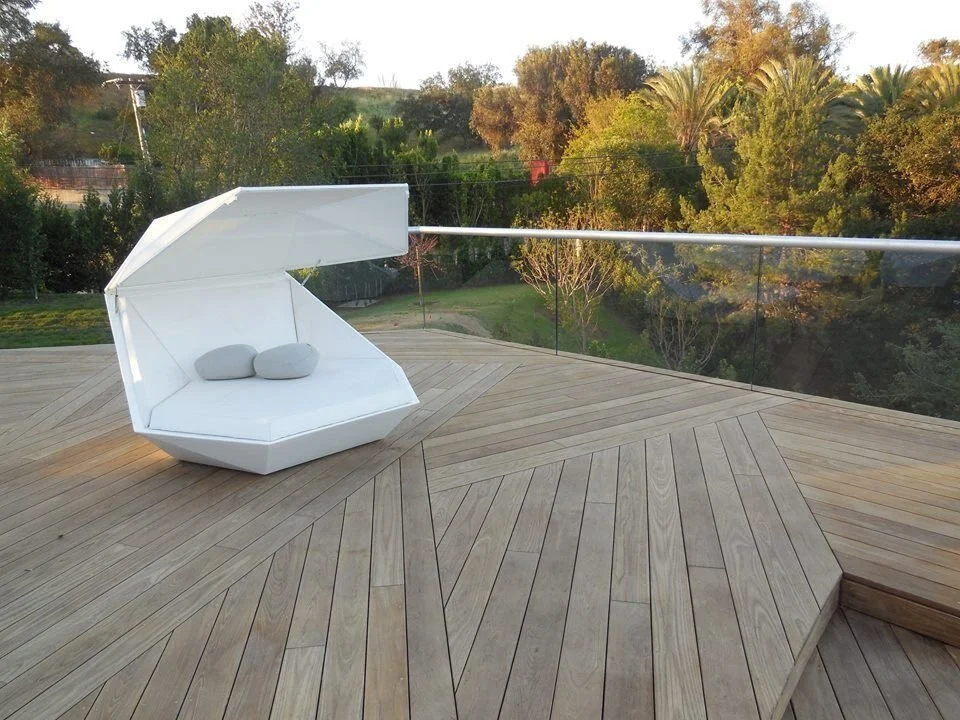 Modern outdoor lounge chair on wooden deck with glass railing and trees in the background.