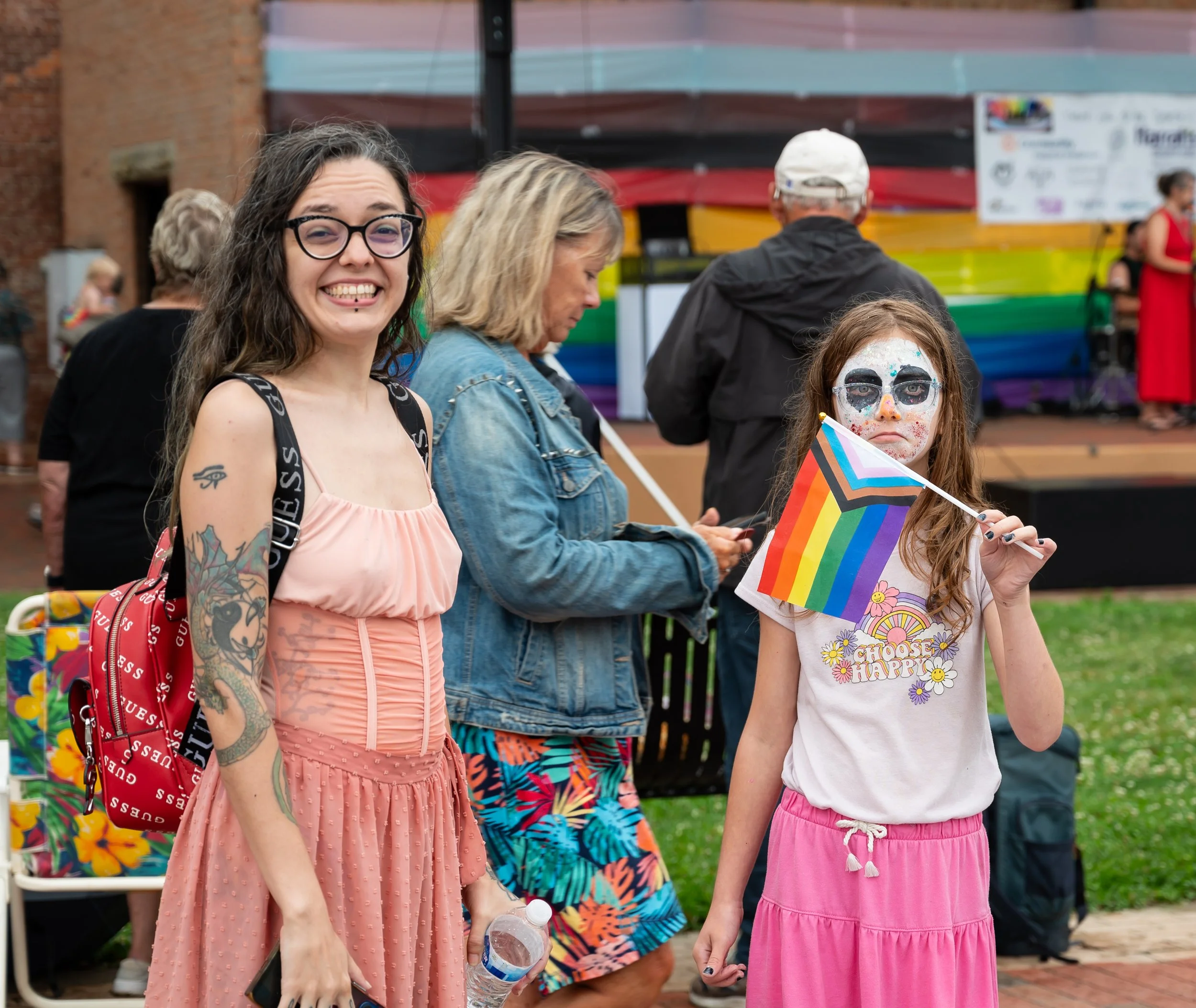 A group of people at an outdoor event; a woman with tattoos and glasses is smiling and holding a water bottle, and a girl with face paint and a rainbow pride flag is looking at the camera.