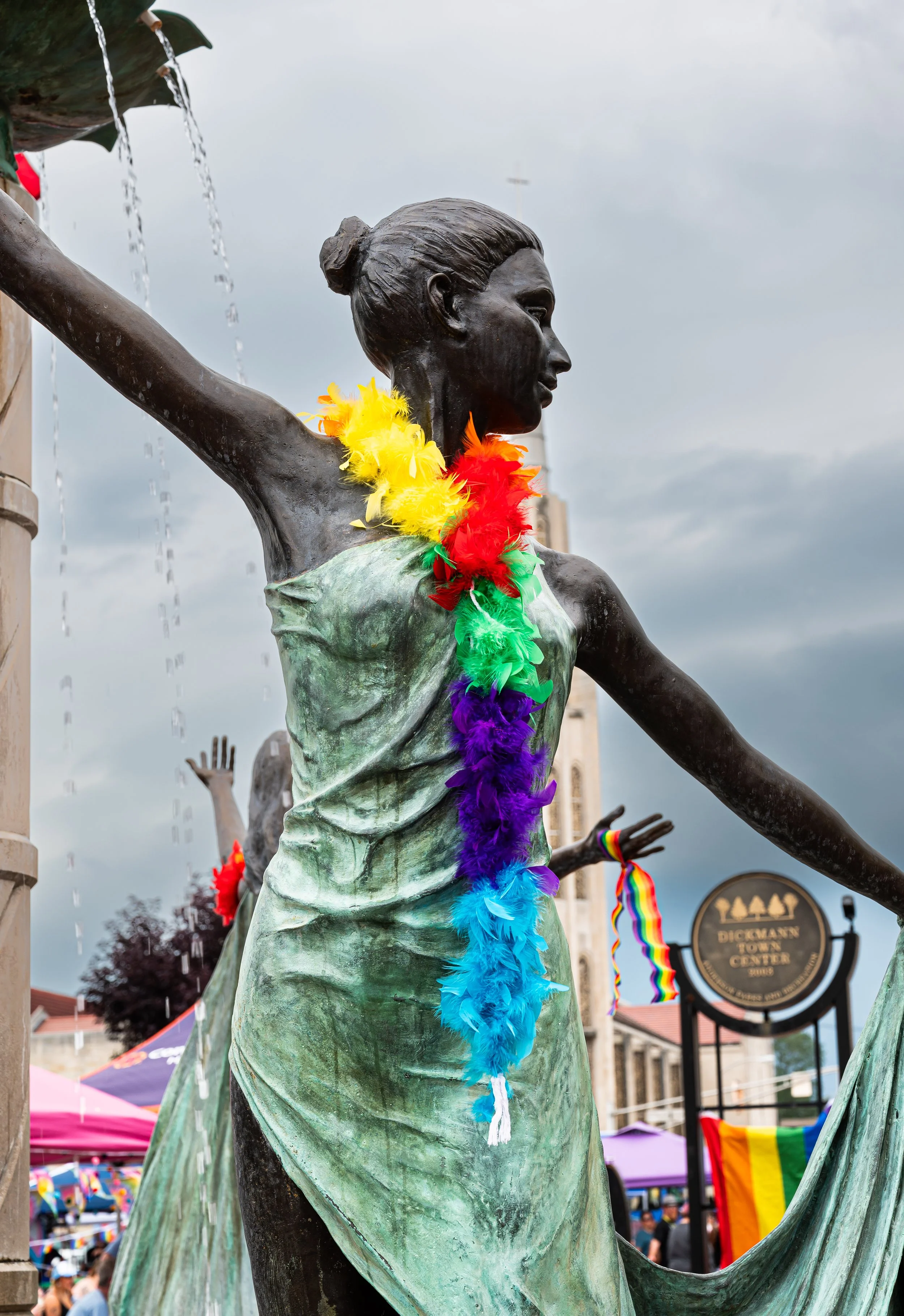 A statue of a woman with a rainbow-colored feather necklace, part of a pride celebration in front of a city hall.