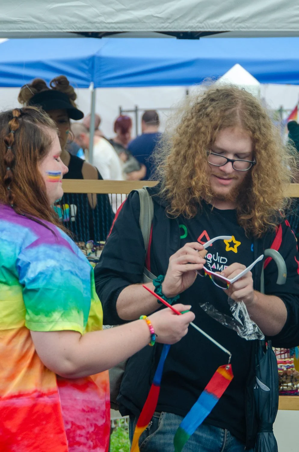 Two people at a pride event, one wearing a rainbow tie-dye shirt with rainbow face paint, the other with curly hair, glasses, and a black jacket, both holding colorful ribbons.