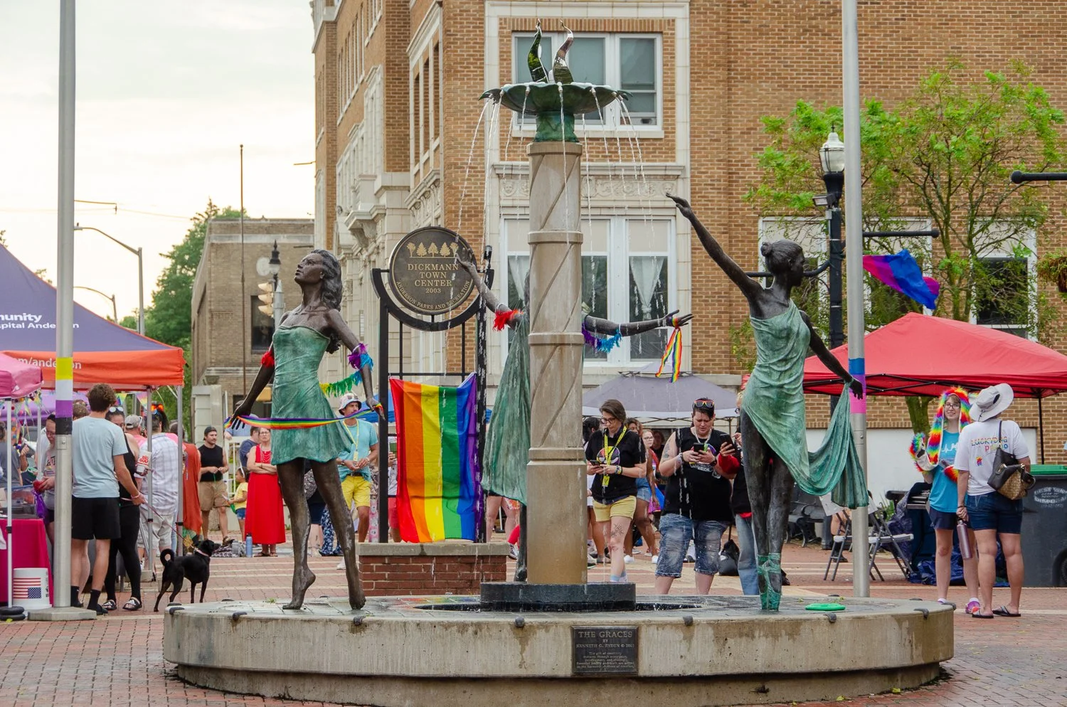 Public event in a city square with a fountain called 'The Graces' featuring three statues of women, decorated with rainbow-colored ribbons and pride flags, surrounded by tents and people celebrating at a pride event.